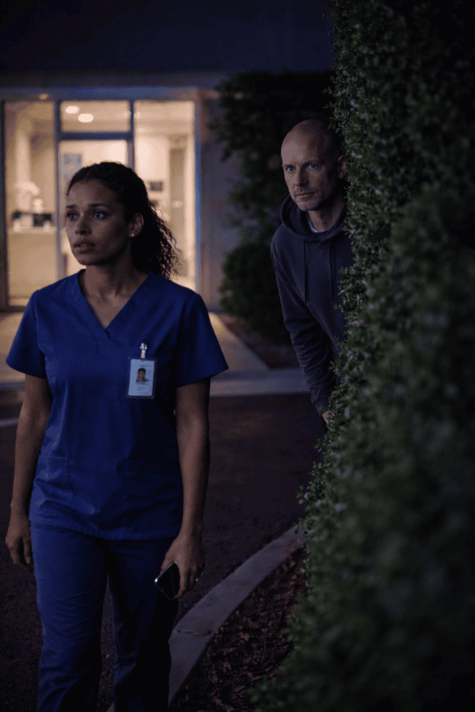Healthcare worker in blue scrubs walking alone at night outside a clinic while a man watches from behind a hedge, illustrating personal safety risks for healthcare professionals