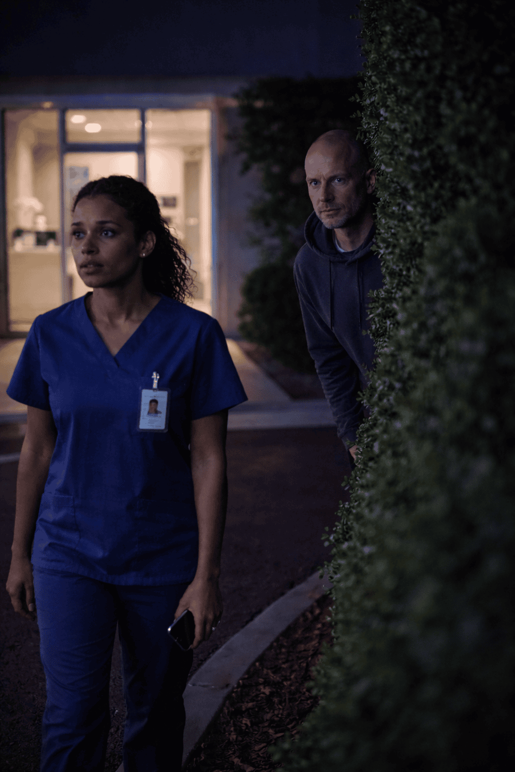 Healthcare worker in blue scrubs walking alone at night outside a clinic while a man watches from behind a hedge, illustrating personal safety risks for healthcare professionals